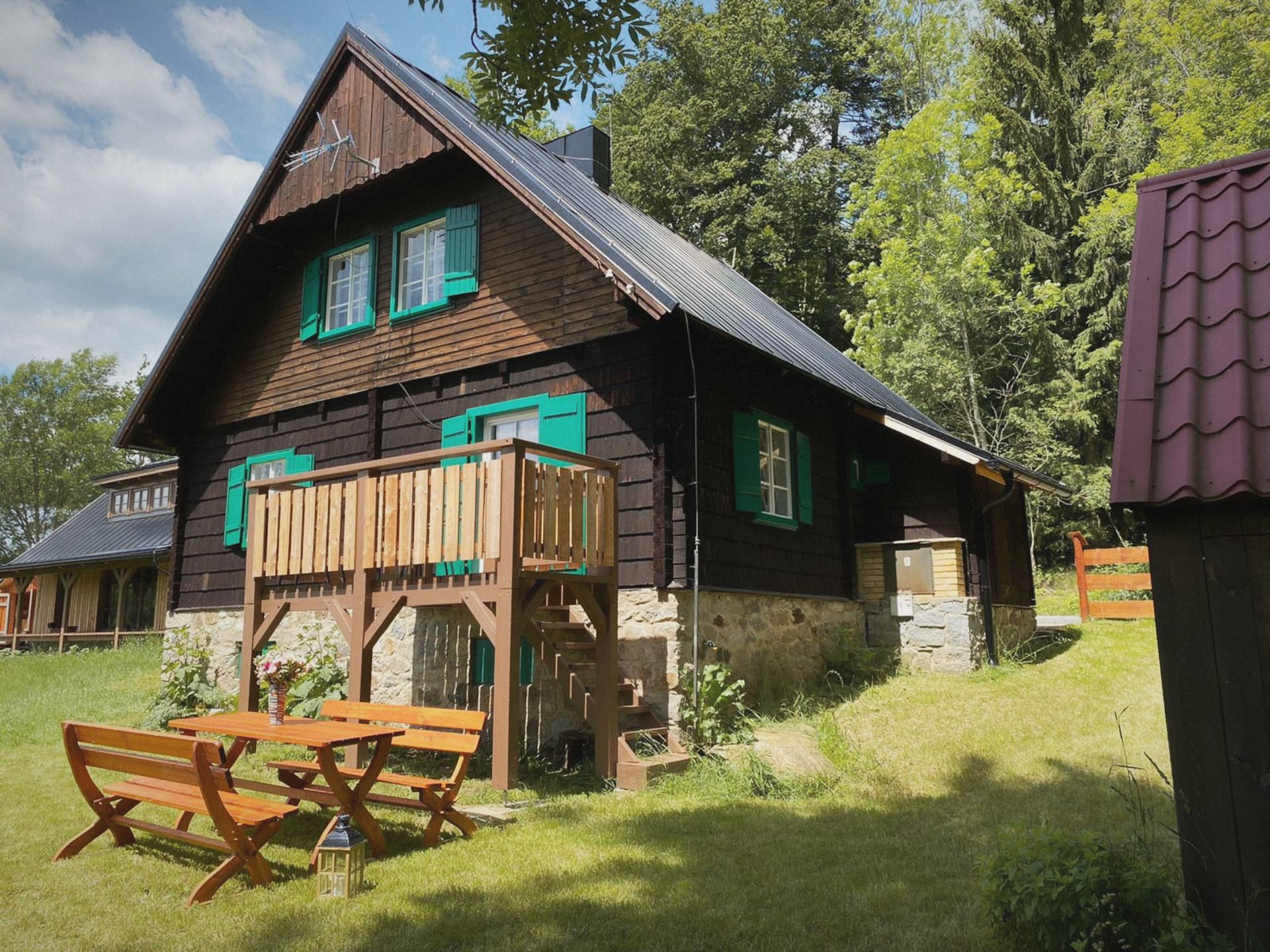 Tmavá dřevěná chata se zelenými okenicemi, balkonem a lavicemi na trávníku u lesa