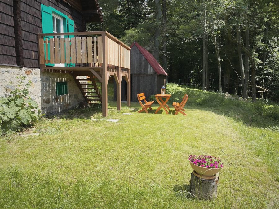 Trávník u chaty s dřevěným balkonem, stolkem a židlemi, za ním les